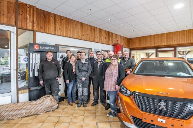 Groupe de personnes posant devant une Peugeot orange dans une concession automobile. Photo d'intérieur avec fond en bois.