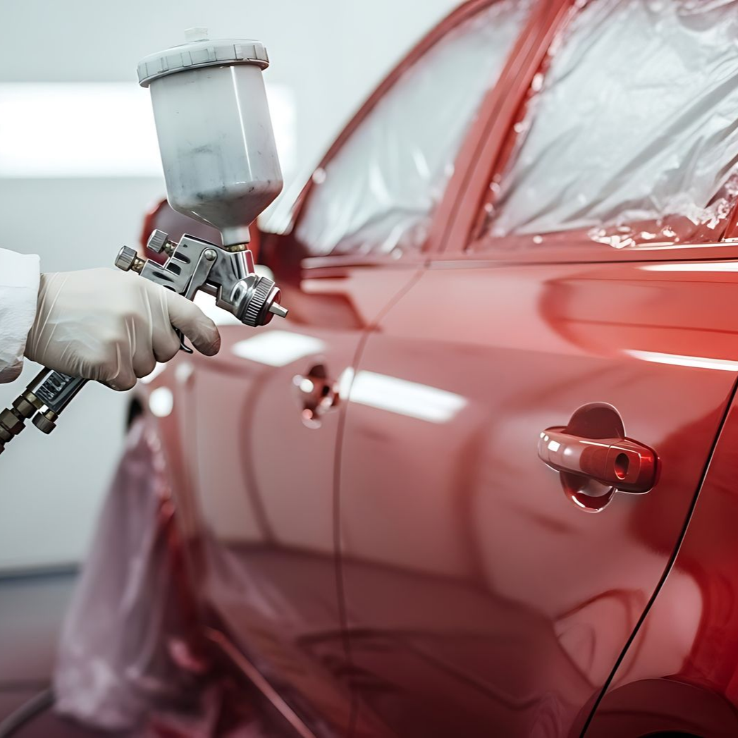 Une personne avec un équipement de protection pulvérise de la peinture rouge sur une voiture dans une cabine de peinture.
