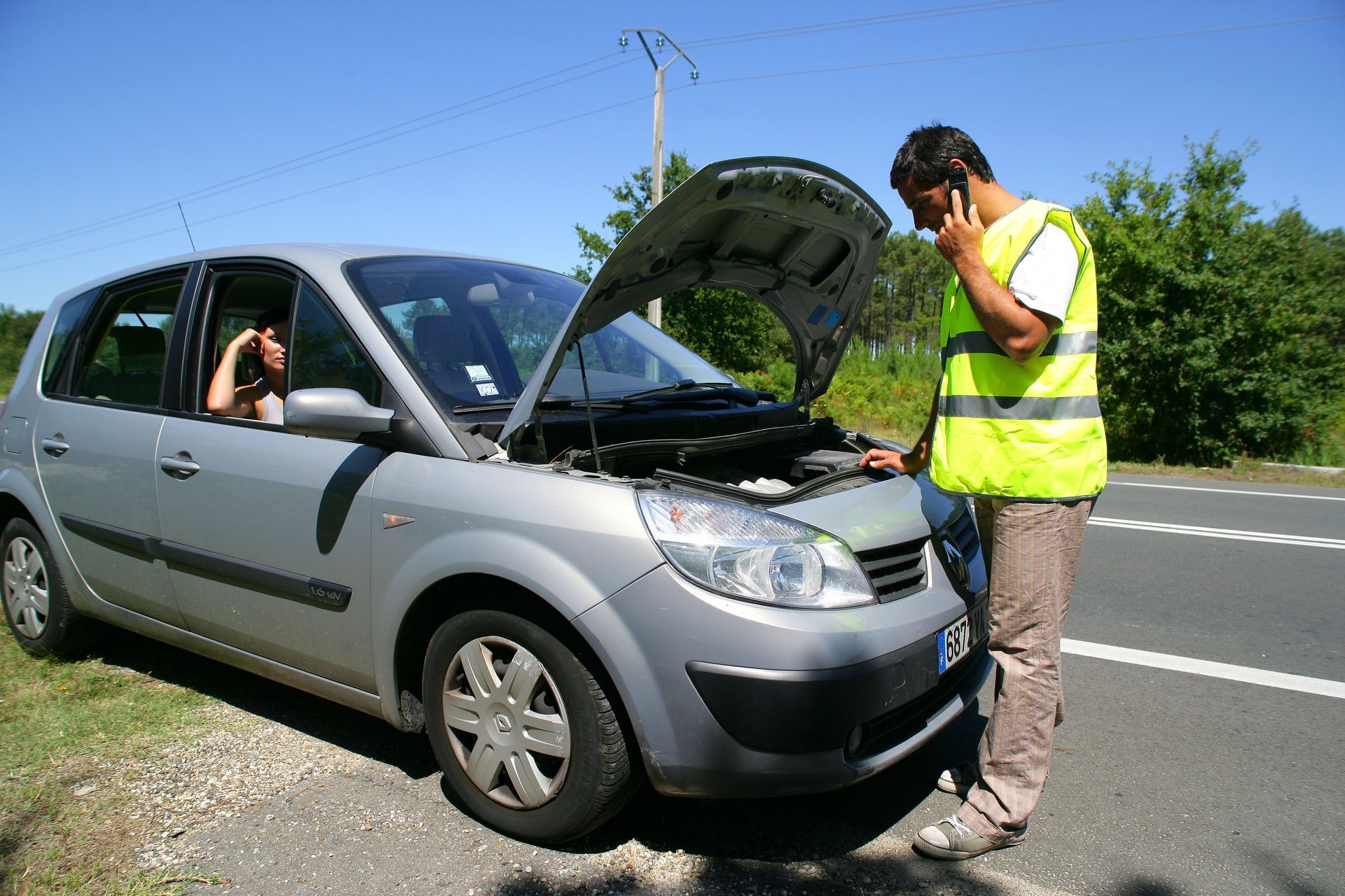 Un homme, capot ouvert, est garé au bord de la route et parle au téléphone ; un passager se trouve à l'intérieur.