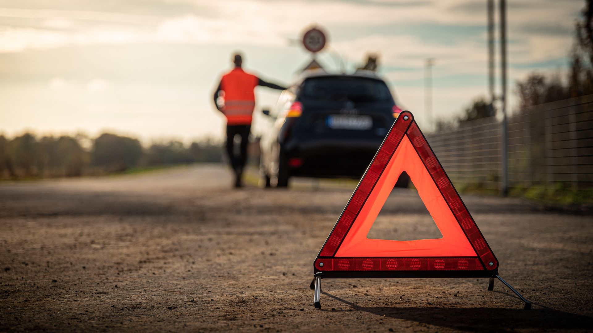 Triangle de signalisation rouge sur la route avec une voiture et une personne portant un gilet orange.