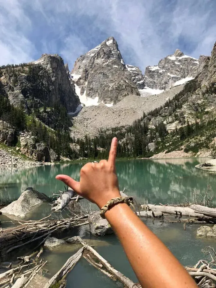 Uma pessoa a fazer um sinal de mão em frente a um lago na montanha.