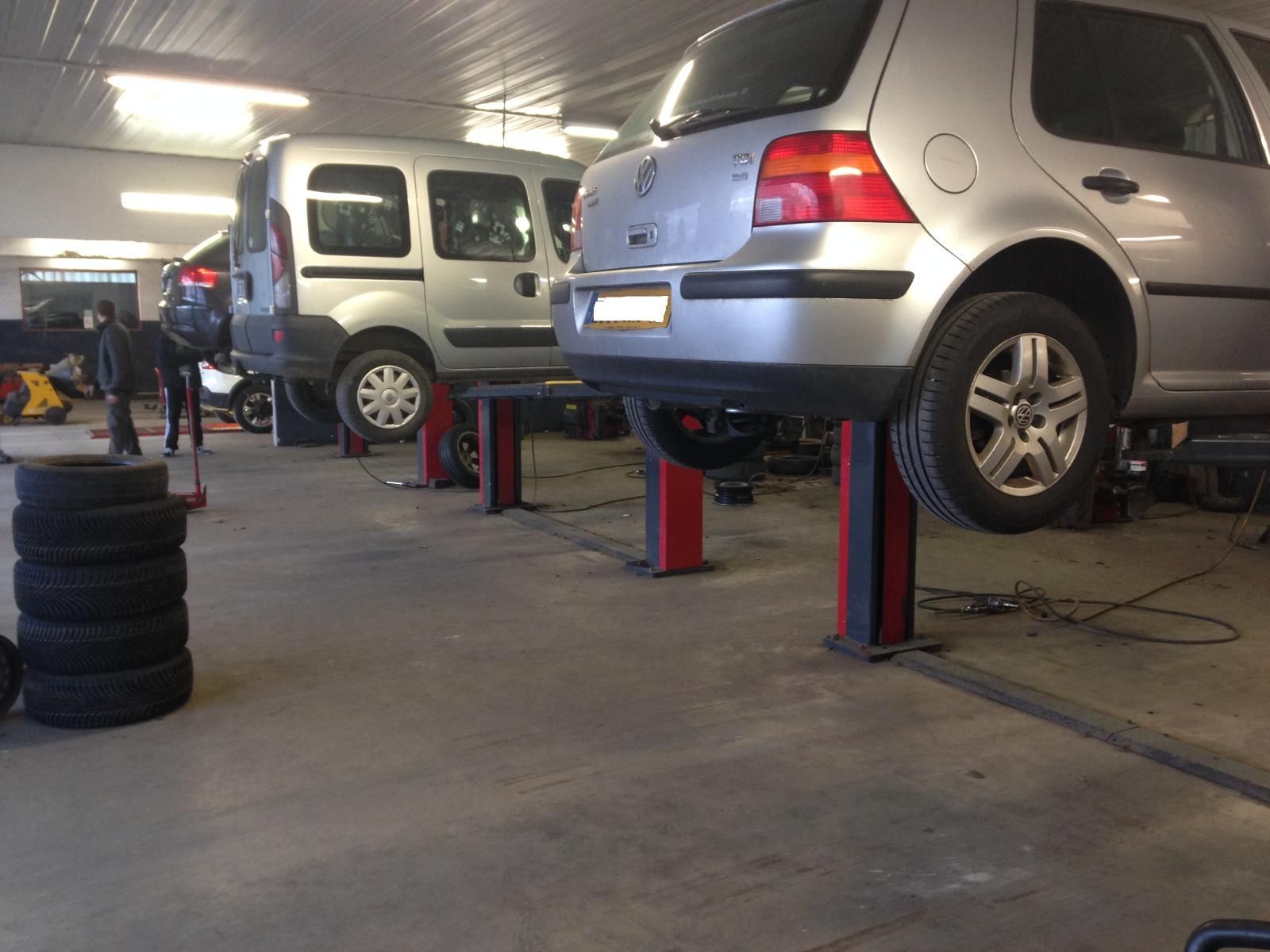 Des voitures sur des ponts élévateurs dans un garage ; des pneus empilés, des outils et une camionnette visibles.