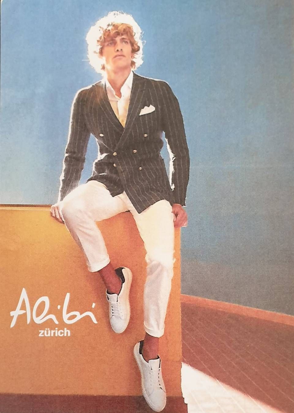 Mann in Nadelstreifenjacke, weisser Hose und Turnschuhen, sitzt auf einer Kiste; Alibi Zürich Logo.