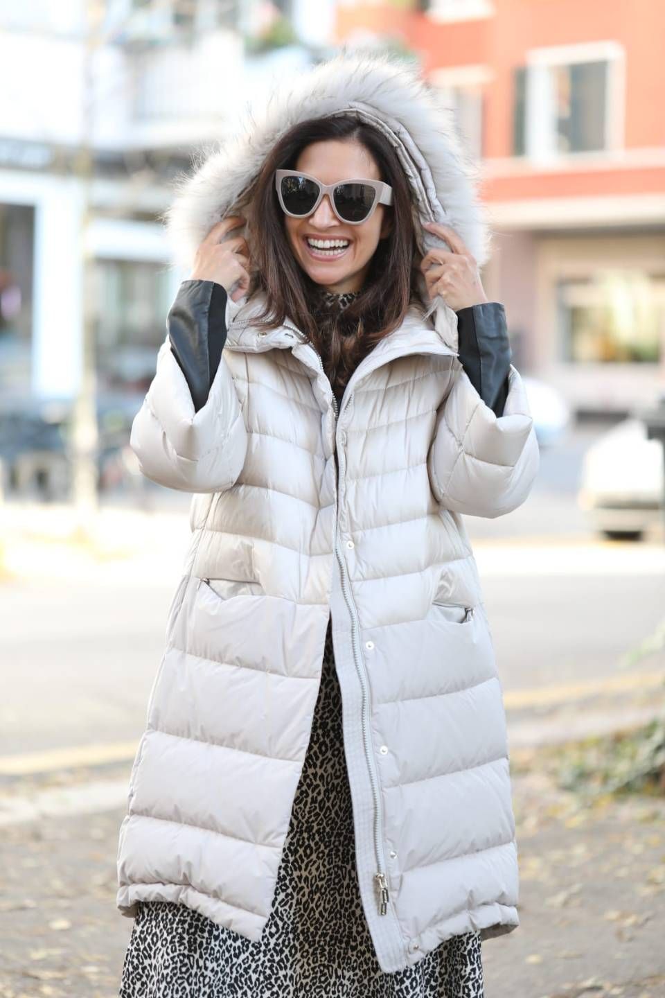 Frau in einem langen, cremefarbenen Daunenmantel mit pelzgefütterter Kapuze, die eine Sonnenbrille trägt, lächelt im Freien.