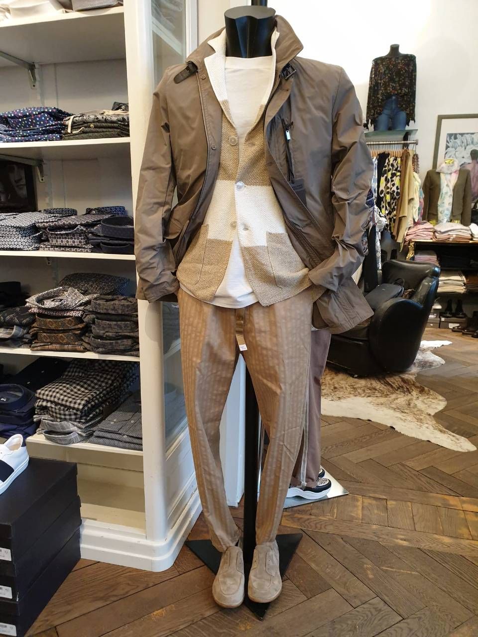 Schaufensterpuppe in mehrlagiger Kleidung: braune Jacke, Weste, Hose und Stiefel. Ladenszene mit Kleiderregalen.