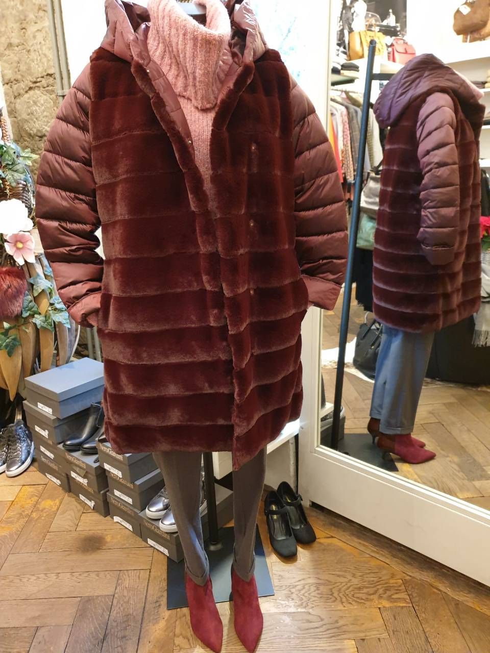 Schaufensterpuppe in einem Geschäft, die einen bordeauxroten Kunstpelzmantel, einen rosa Pullover und rote Stiefel trägt.