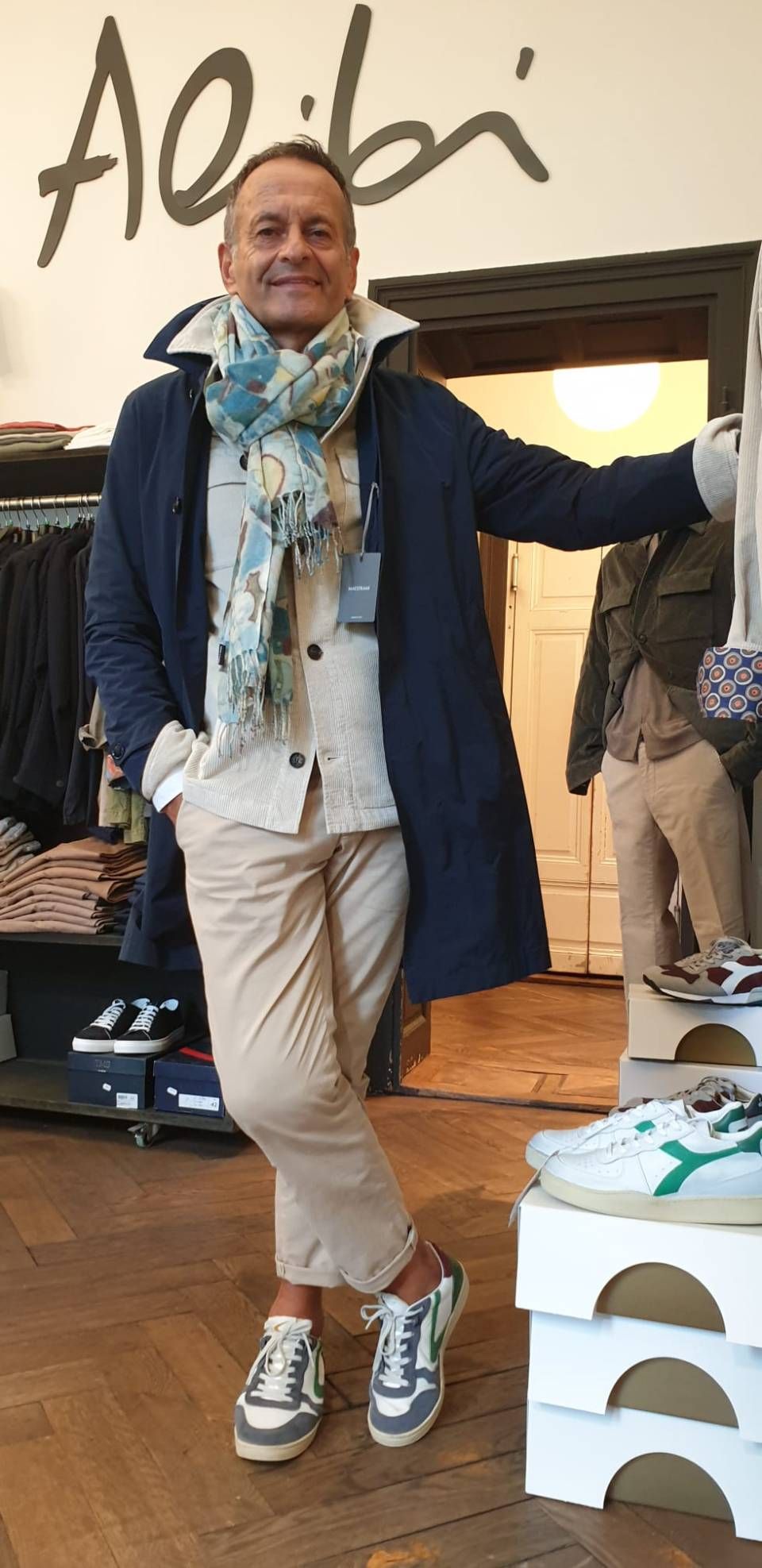 Ein Mann bekleidet mit Mantel, Schal und Turnschuhen. Er lächelt. Er lehnt an der Theke neben gestapelten Schuhkartons.