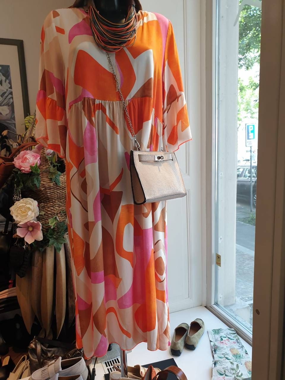 Schaufensterpuppe in einem leuchtend orange-pink gemusterten Kleid mit Handtasche und Halskette als Accessoires.