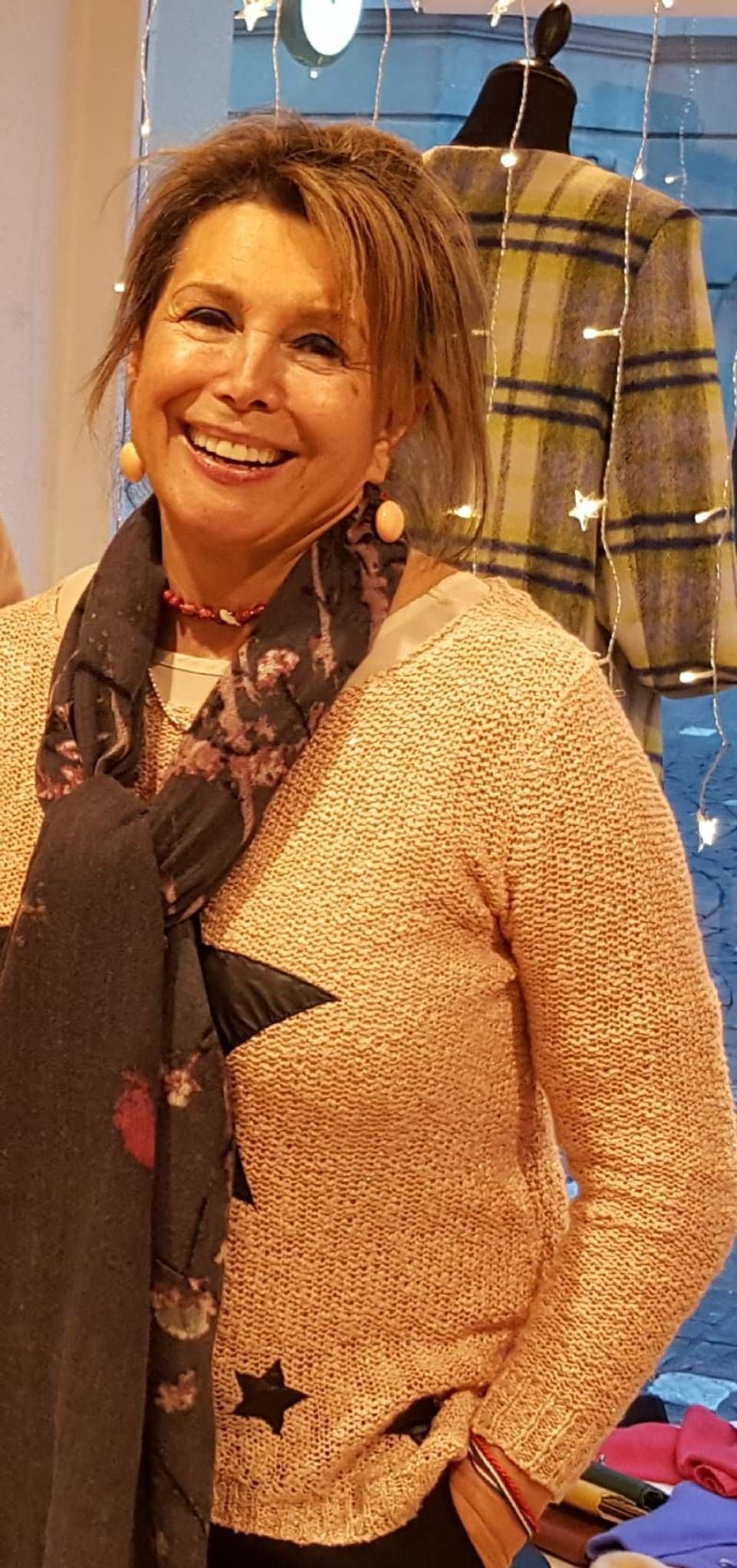 Eine Frau mit einem Schal und einem pfirsichfarbenen Pullover lächelt den Betrachter an; sie steht in einem Geschäft.