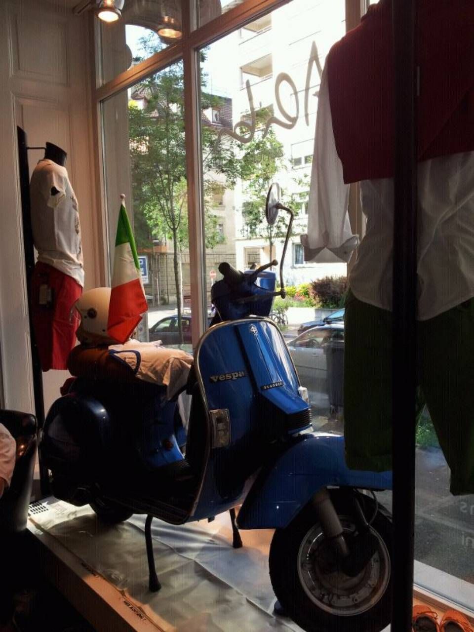Blauer Vespa-Roller mit italienischer Flagge, geparkt in einem Schaufenster neben Kleidung.