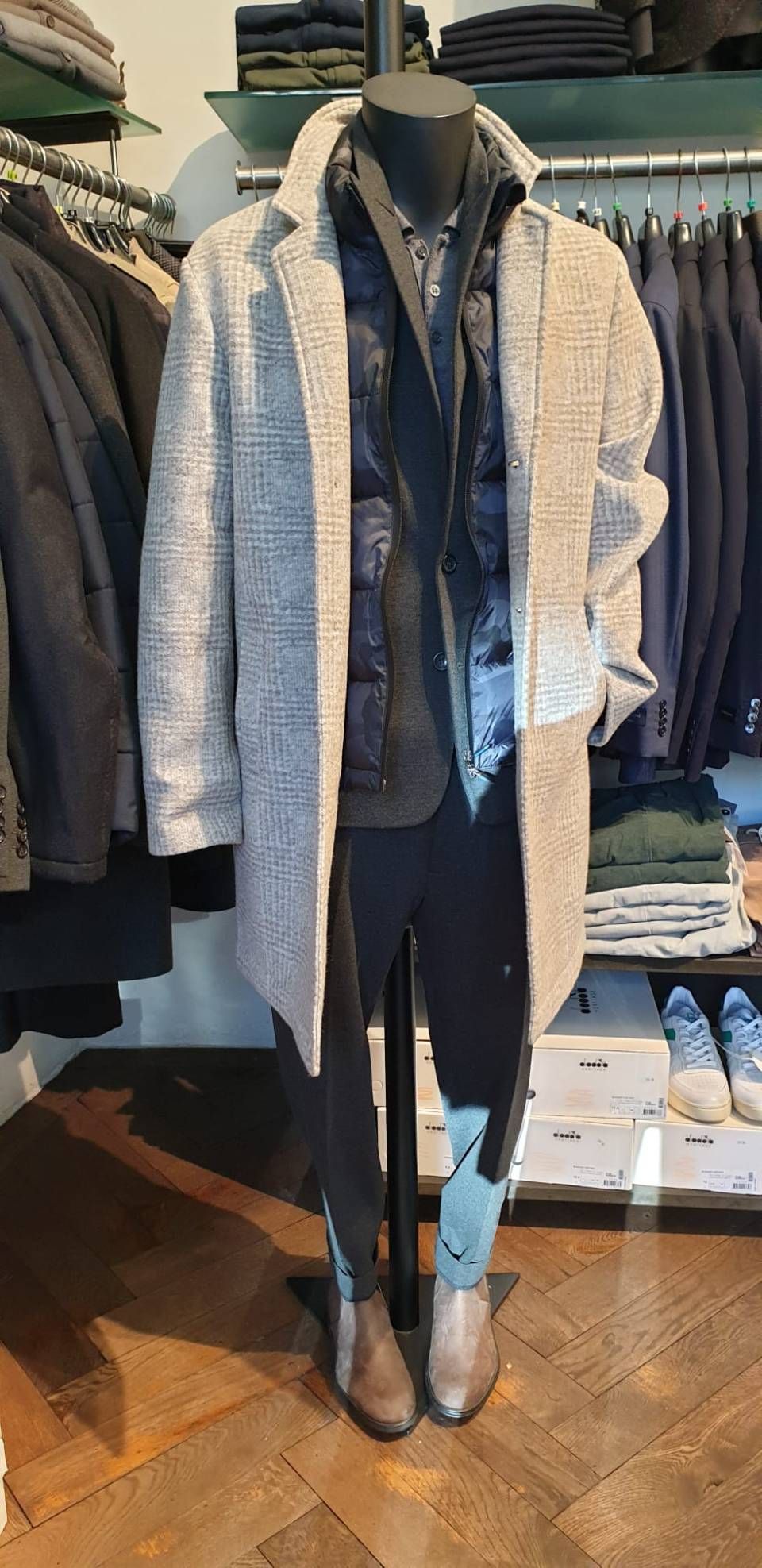 Eine Schaufensterpuppe trägt einen grauen Mantel, eine schwarze Steppweste, ein Hemd, eine schwarze Hose braune Stiefel.