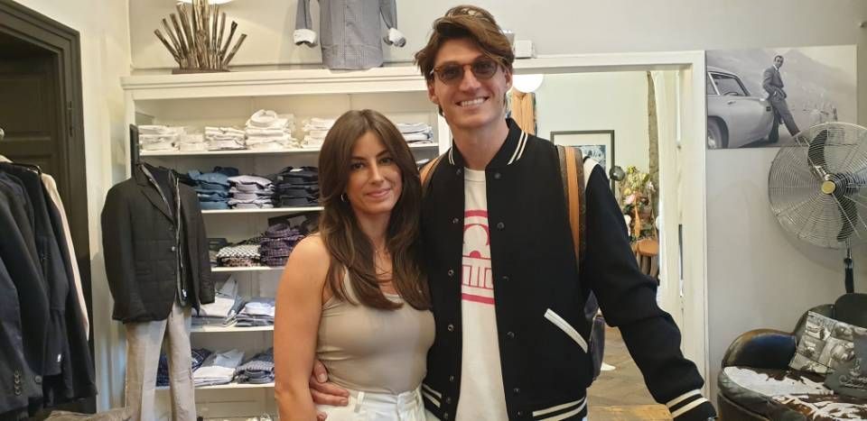 Ein Paar in einem Bekleidungsgeschäft. Der Mann trägt eine Sonnenbrille und eine Collegejacke. Die Frau trägt ein Tanktop.