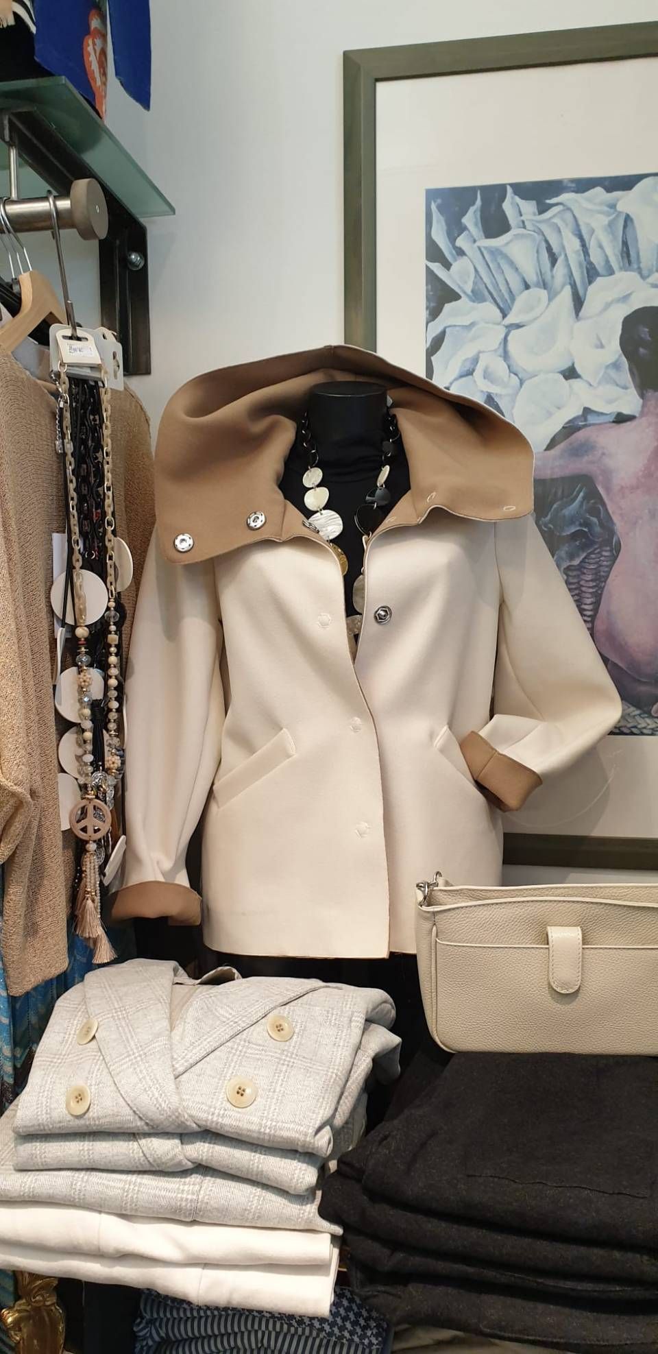 Eine Schaufensterpuppe trägt eine cremefarbene Jacke mit hellbraunem Kragen. Accessoires und gefaltete Kleidung.