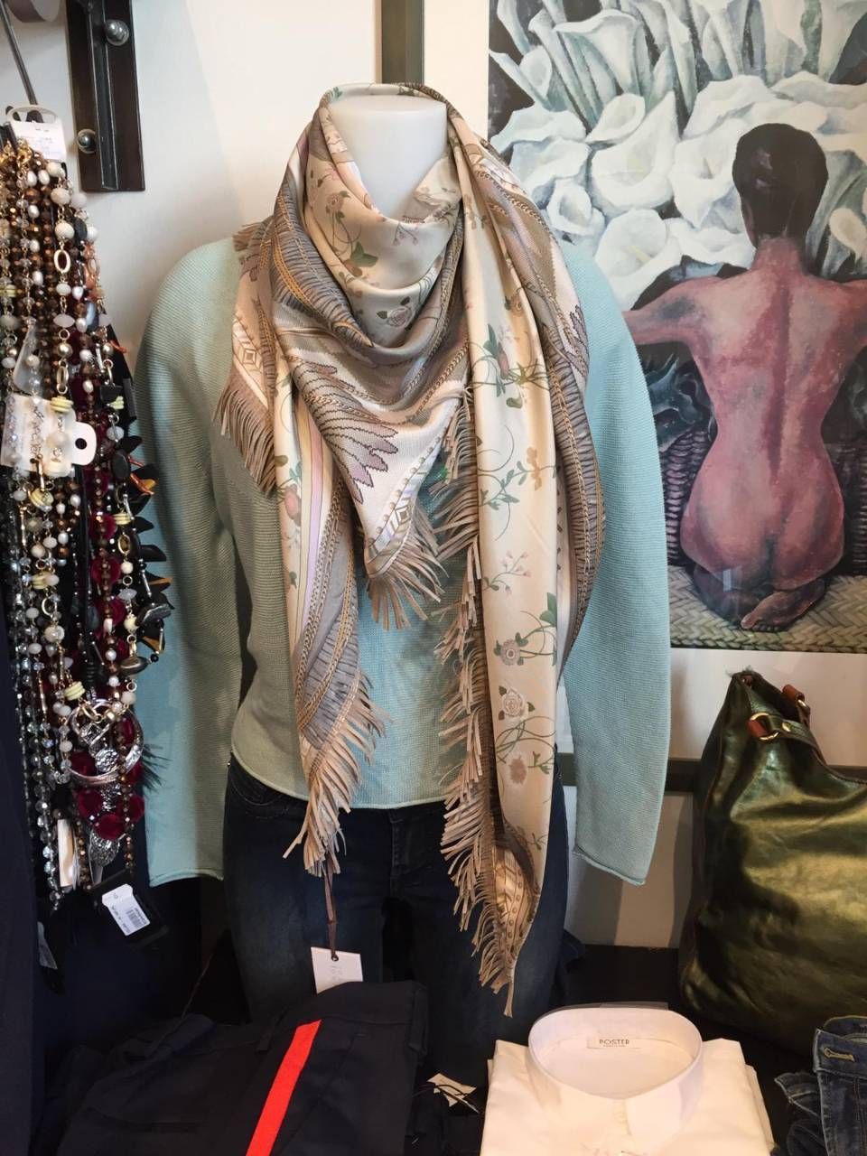 Eine Schaufensterpuppe trägt einen Schal, einen Pullover und Jeans; im Hintergrund sind Kunstwerke zu sehen.