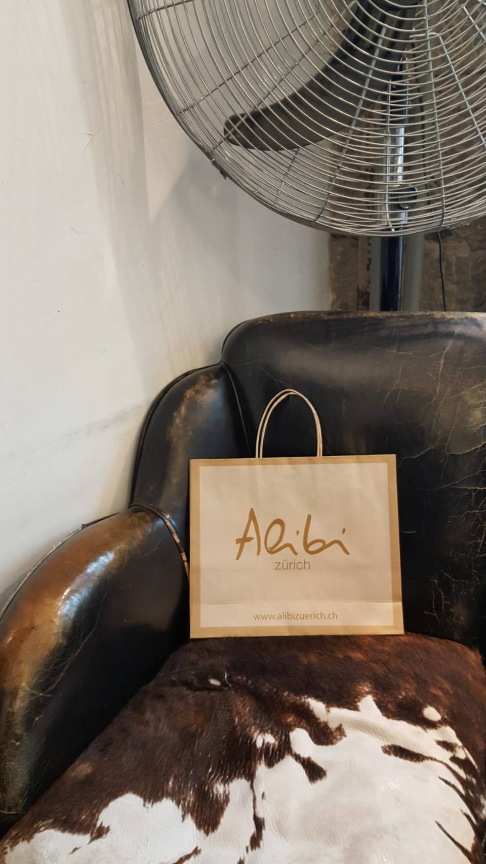 Auf einem mit Kuhfell bezogenen Stuhl neben einem Ventilator steht eine braune Papiertüte mit der Aufschrift „Alibi“.