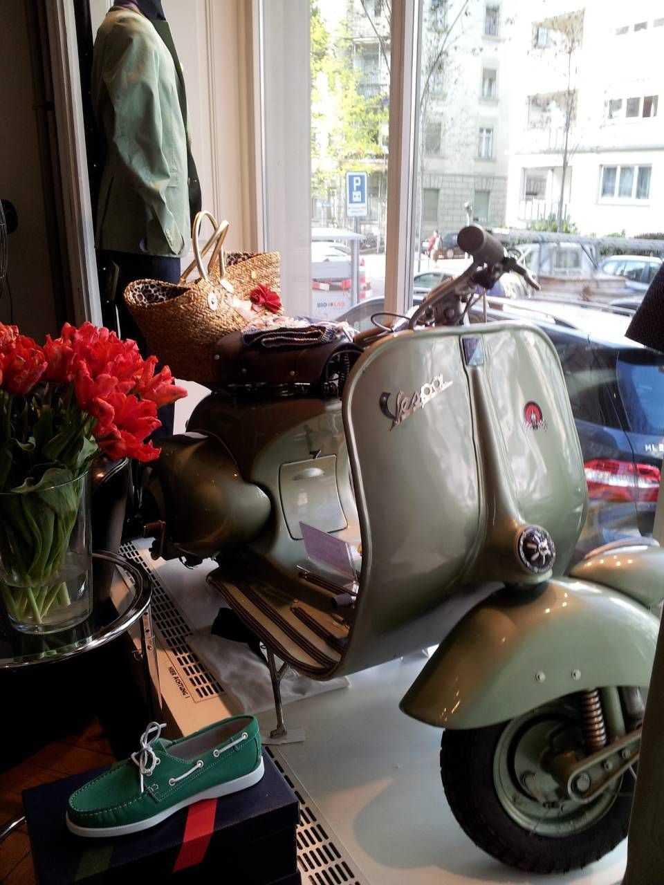 Ein grüner Vespa-Roller steht in einem Schaufenster neben Schuhen, Blumen und einer Strohtasche.
