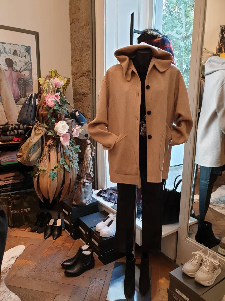 Schaufensterpuppe in einem beigen Mantel, schwarzer Hose und Stiefeln, in einem Ladenambiente mit Kleidung und Accessoires.