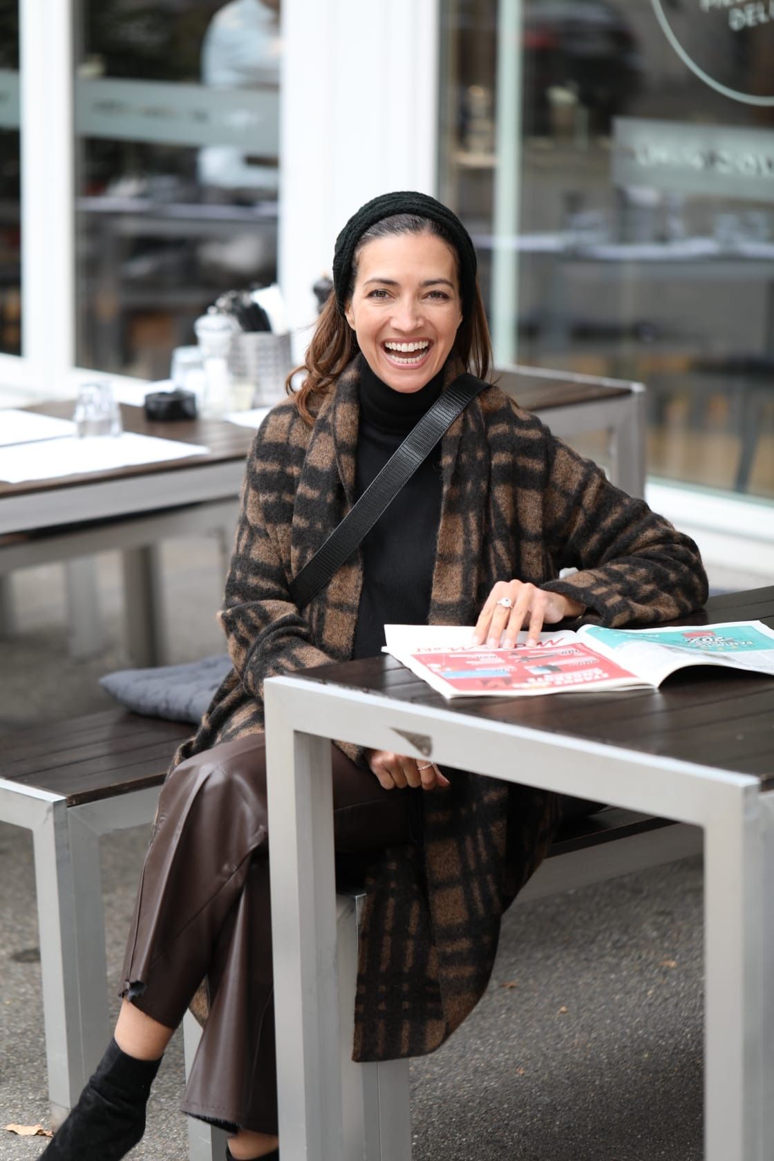 Eine lächelnde Frau sitzt im Freien an einem Tisch, trägt einen Mantel und liest eine Zeitschrift.