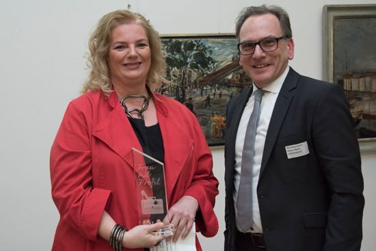 Eine Frau in einer roten Jacke hält eine Auszeichnung neben einem Mann im Anzug