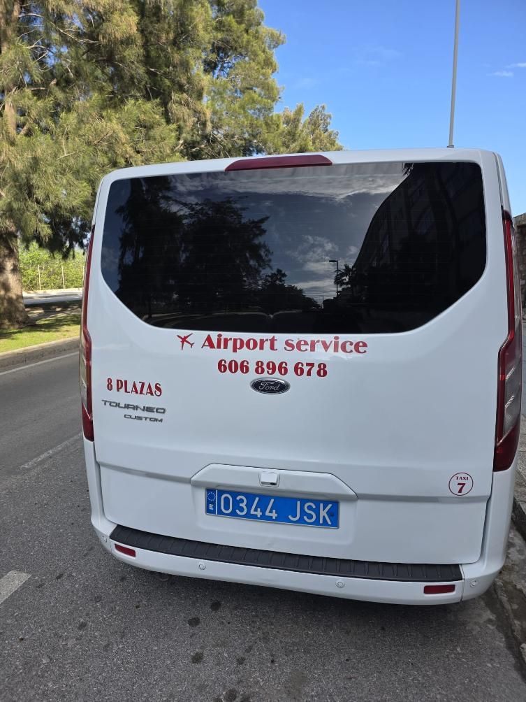 Una furgoneta blanca de transporte al aeropuerto con letras rojas, aparcada al borde de la carretera cerca de unos árboles.