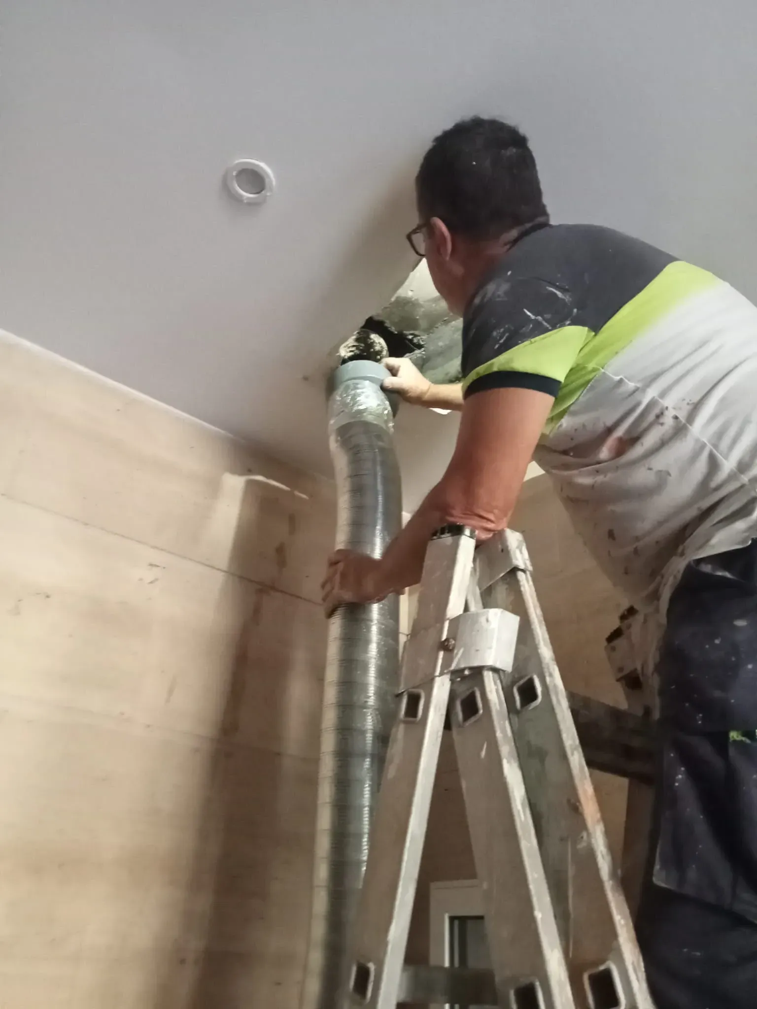 Un trabajador subido a una escalera instala una tubería metálica cerca del techo de una habitación.