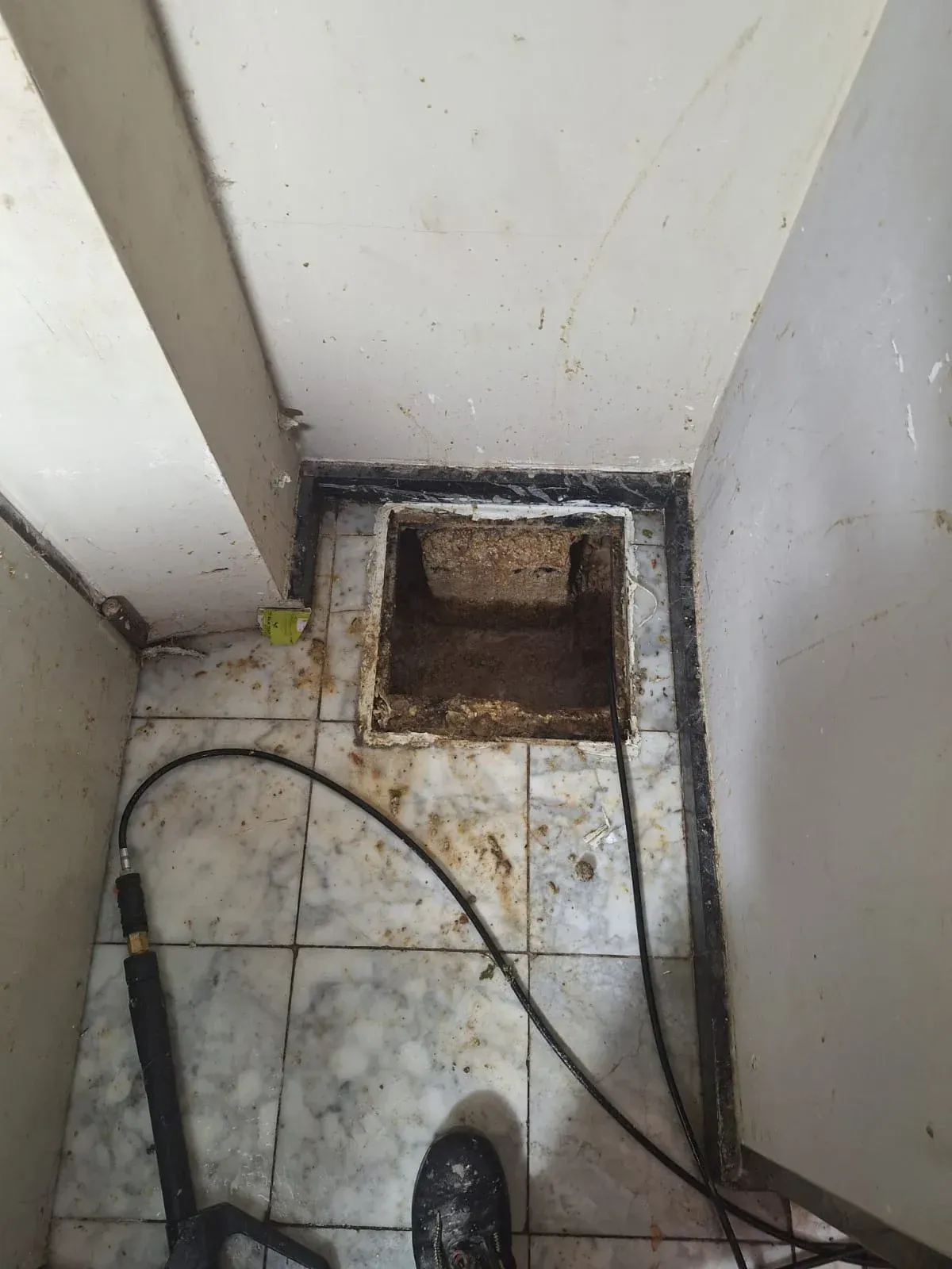Espacio de servicio estrecho con desagüe abierto, suelo de baldosas sucio y cables en el suelo.