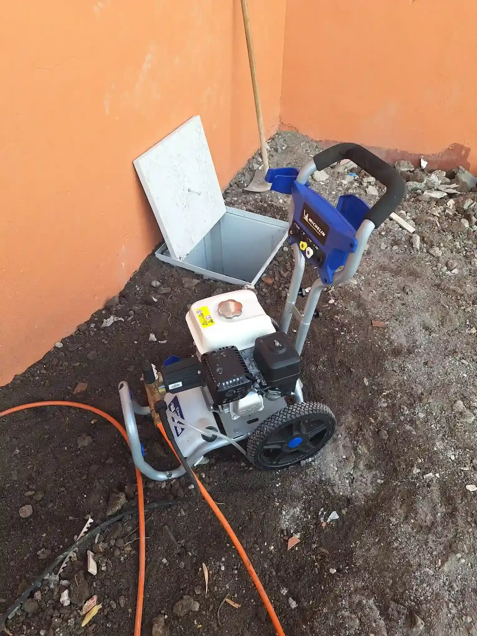 Pequeño robot con ruedas junto a una pared, con una caja abierta y un cable naranja sobre una superficie de tierra y grava.