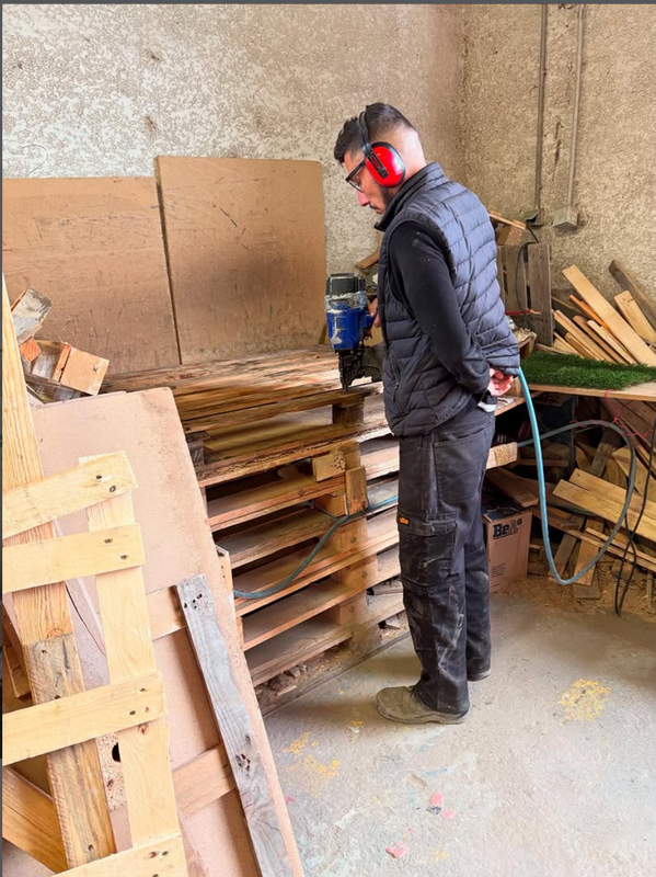 Un ouvrier portant un casque antibruit rouge inspecte des palettes de bois empilées dans un atelier.