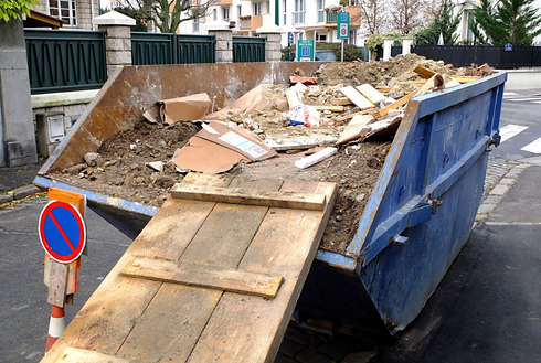 Une benne à ordures bleue débordant de débris de construction se trouve dans une rue, avec une rampe en bois appuyée contre elle.