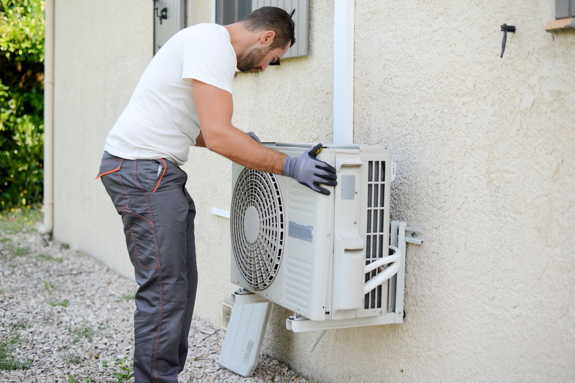 Un homme en vêtements de travail gris installe une unité de climatisation extérieure sur un mur de maison beige.
