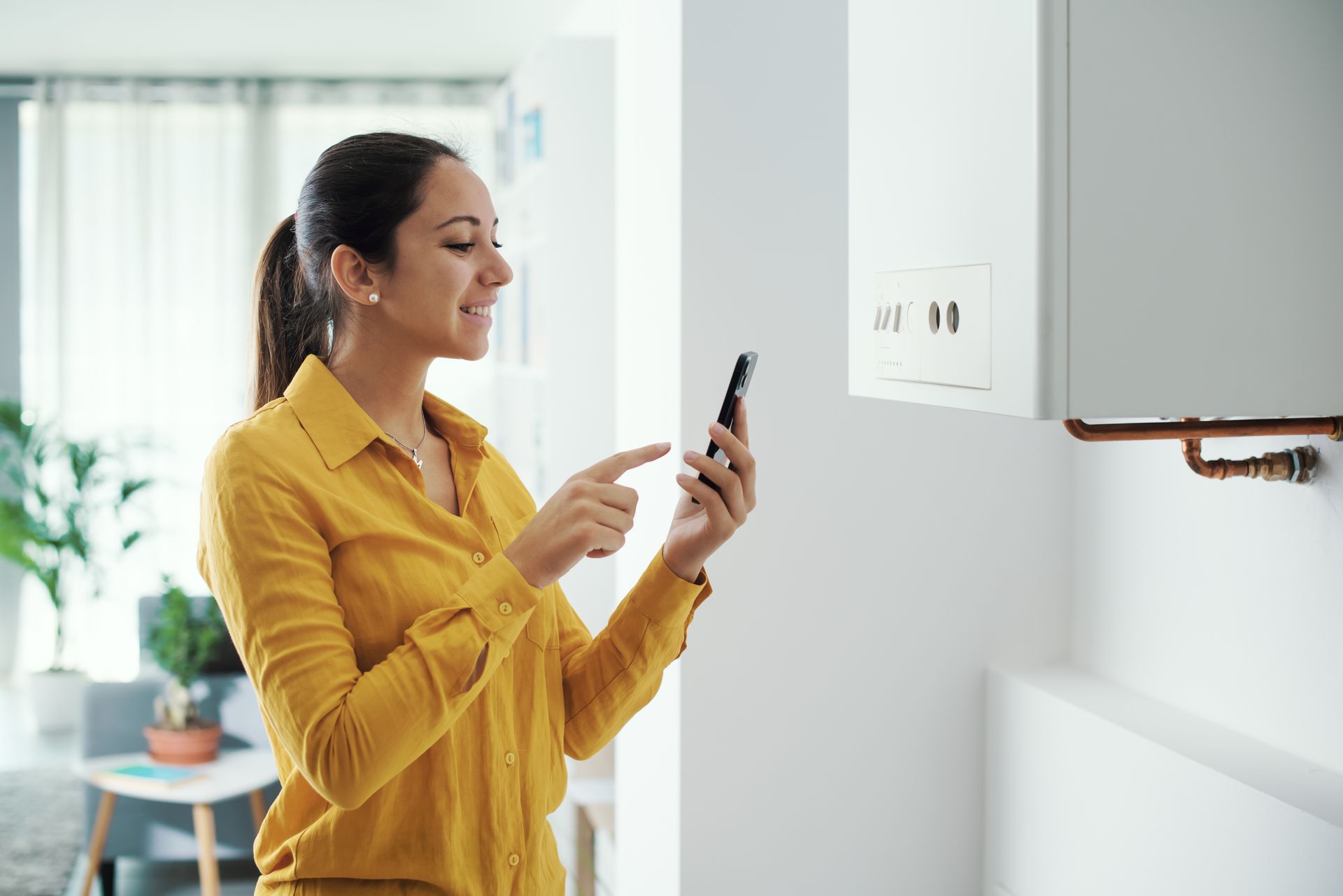 Une femme en chemise jaune sourit, utilisant son smartphone près d'un radiateur mural blanc.