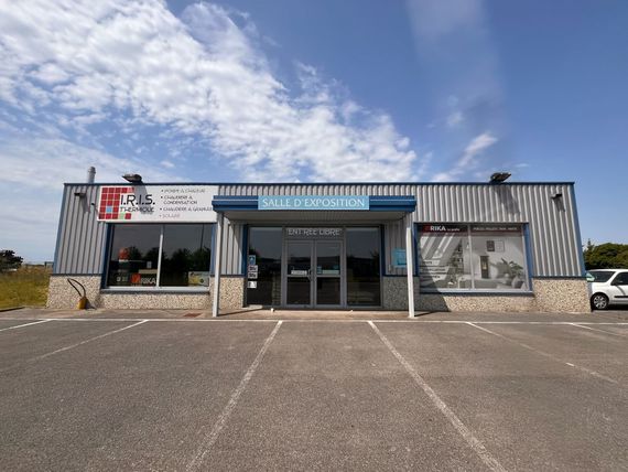 Un bâtiment commercial bleu et gris avec une porte d'entrée en verre et des vitrines, un parking devant.