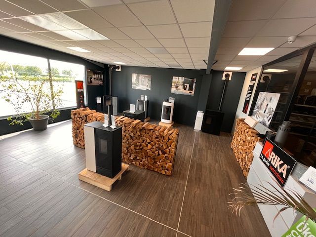 Salle d'exposition avec poêles à bois, présentoir de piles de bois, parquet et grandes fenêtres.