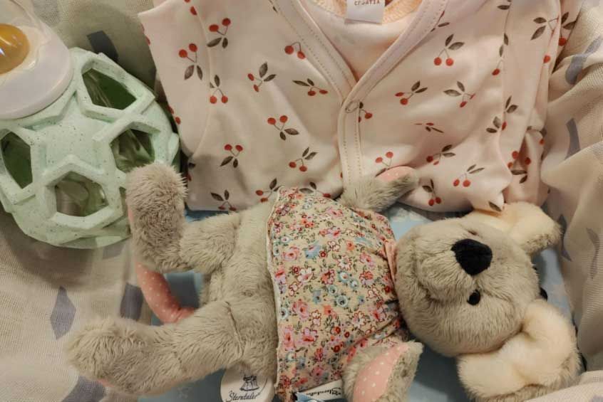Ein Stoffkoala trägt ein Kleid mit Blumenmuster. Dahinter befindet sich ein rosafarbenes Oberteil für Babys mit kleinen Kirschen als Muster.