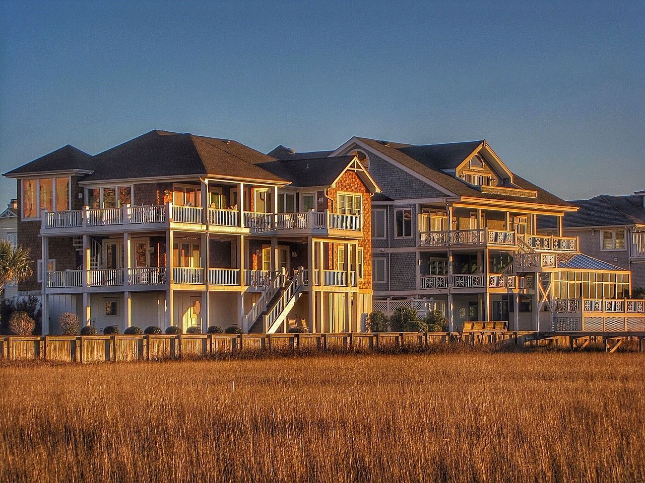 Großes, mehrstöckiges Strandhaus mit Balkonen vor blauem Himmel, gesehen von einem Feld mit hohem, goldenem Gras.