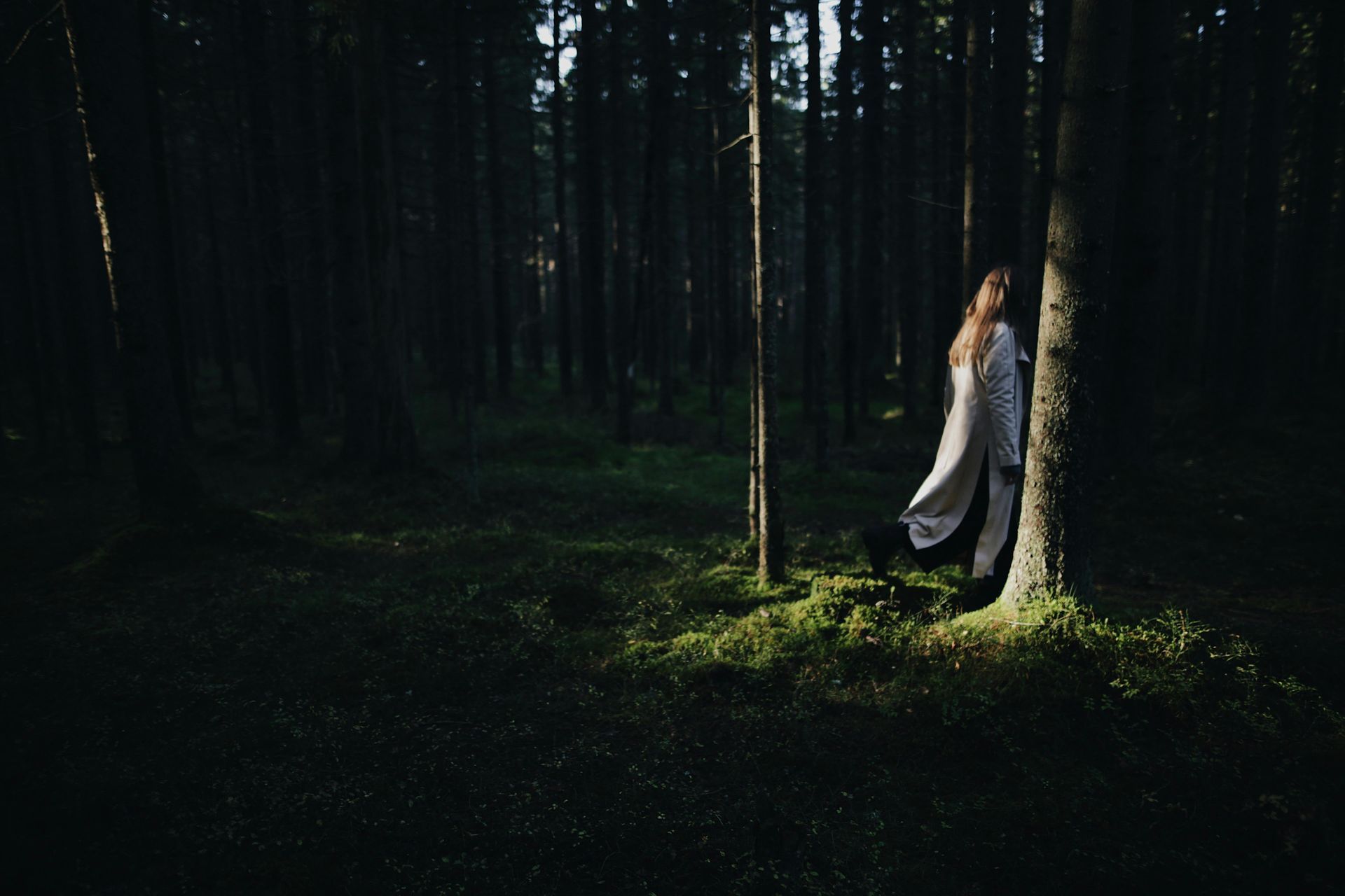 Frau in weißem Kleid lehnt an einem Baum in einem dunklen Wald, Sonnenlicht fällt auf Moos.