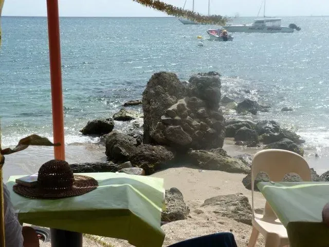 Vue du restaurant en bord de mer : chapeau sur la table, formation rocheuse dans l'eau, bateaux et mer turquoise claire.
