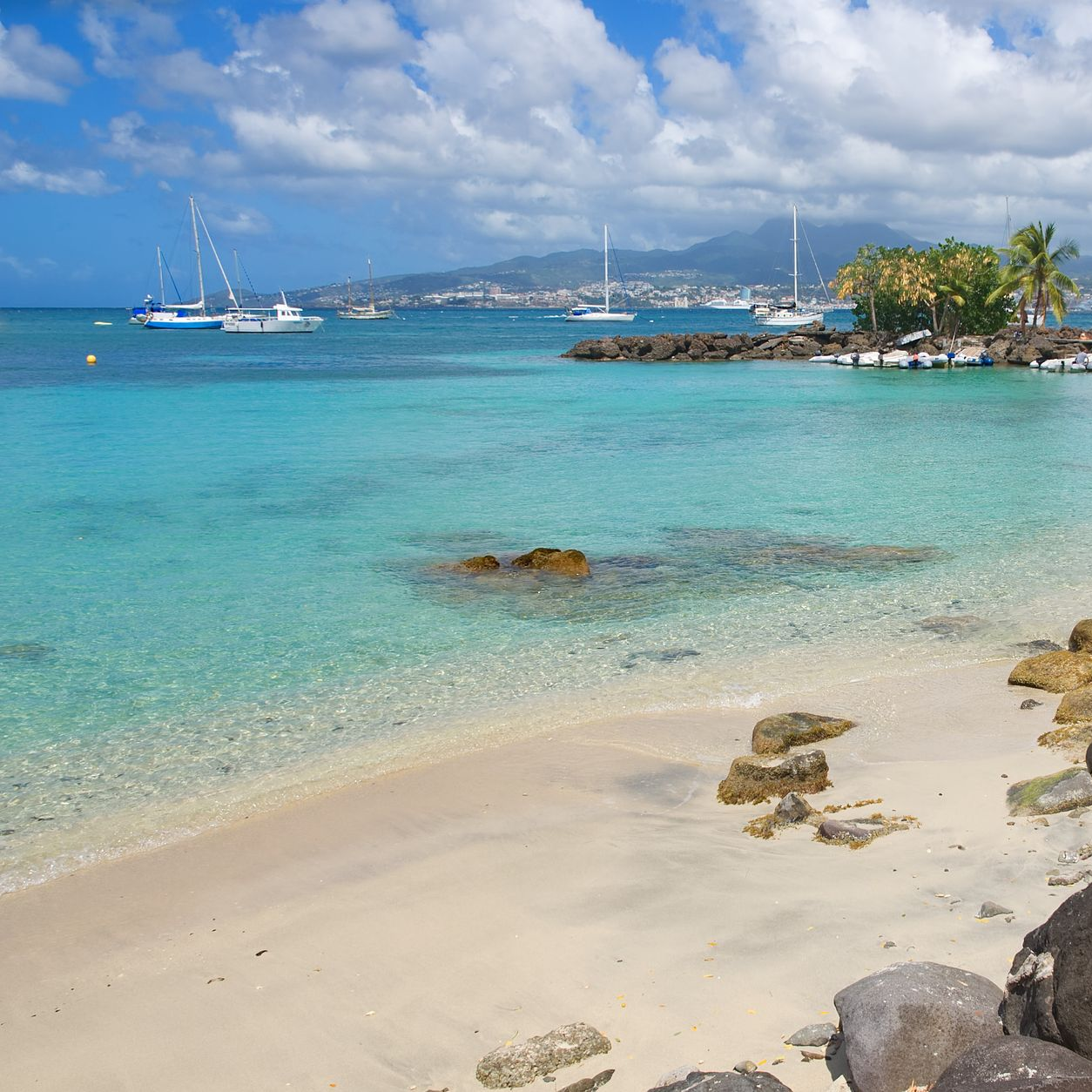 Plage de sable avec eau turquoise claire, voiliers et vue sur une ville lointaine sous un ciel ensoleillé.