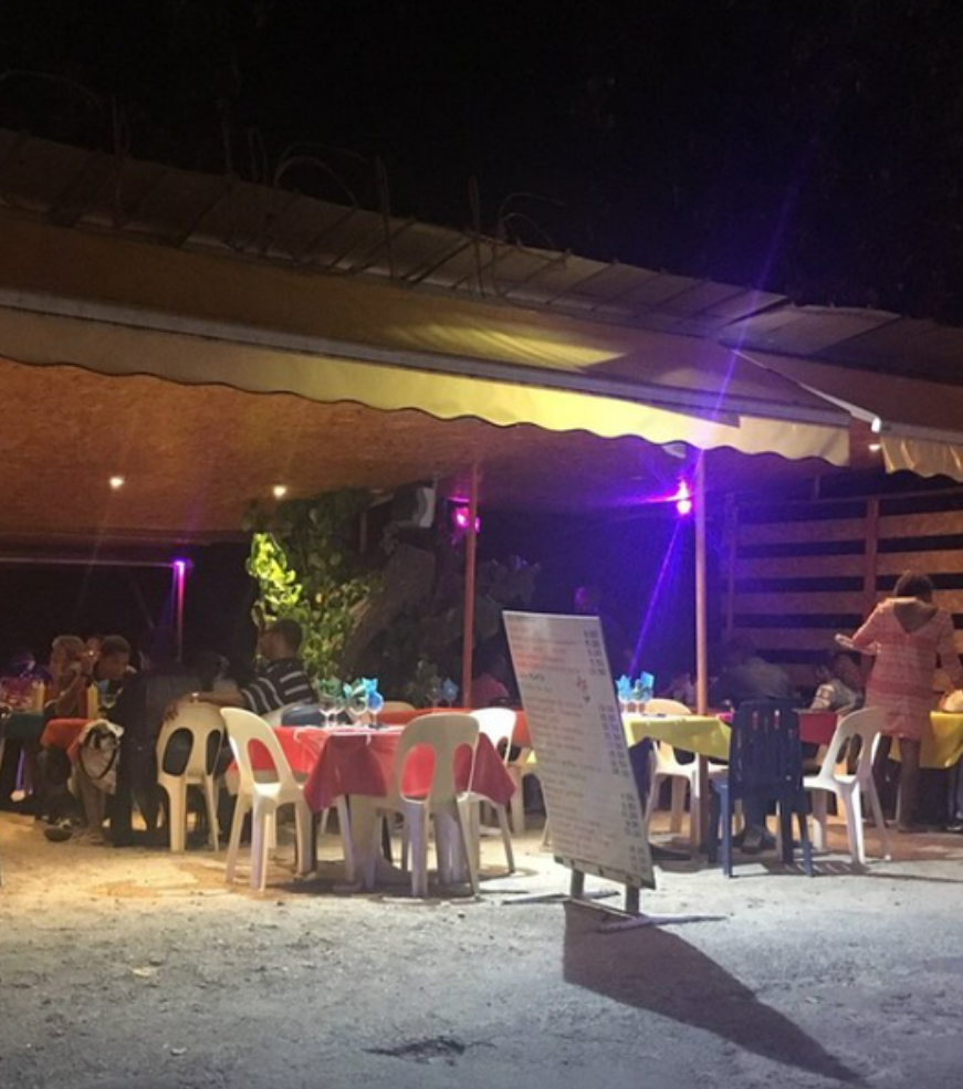 Restaurant en plein air le soir, avec tables, personnes et tableau de menu sous un auvent.