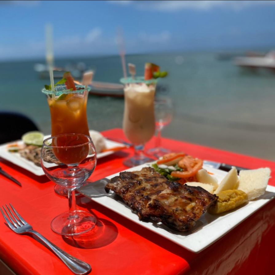 Côtes grillées, cocktails et accompagnements sur une table rouge au bord de la mer.