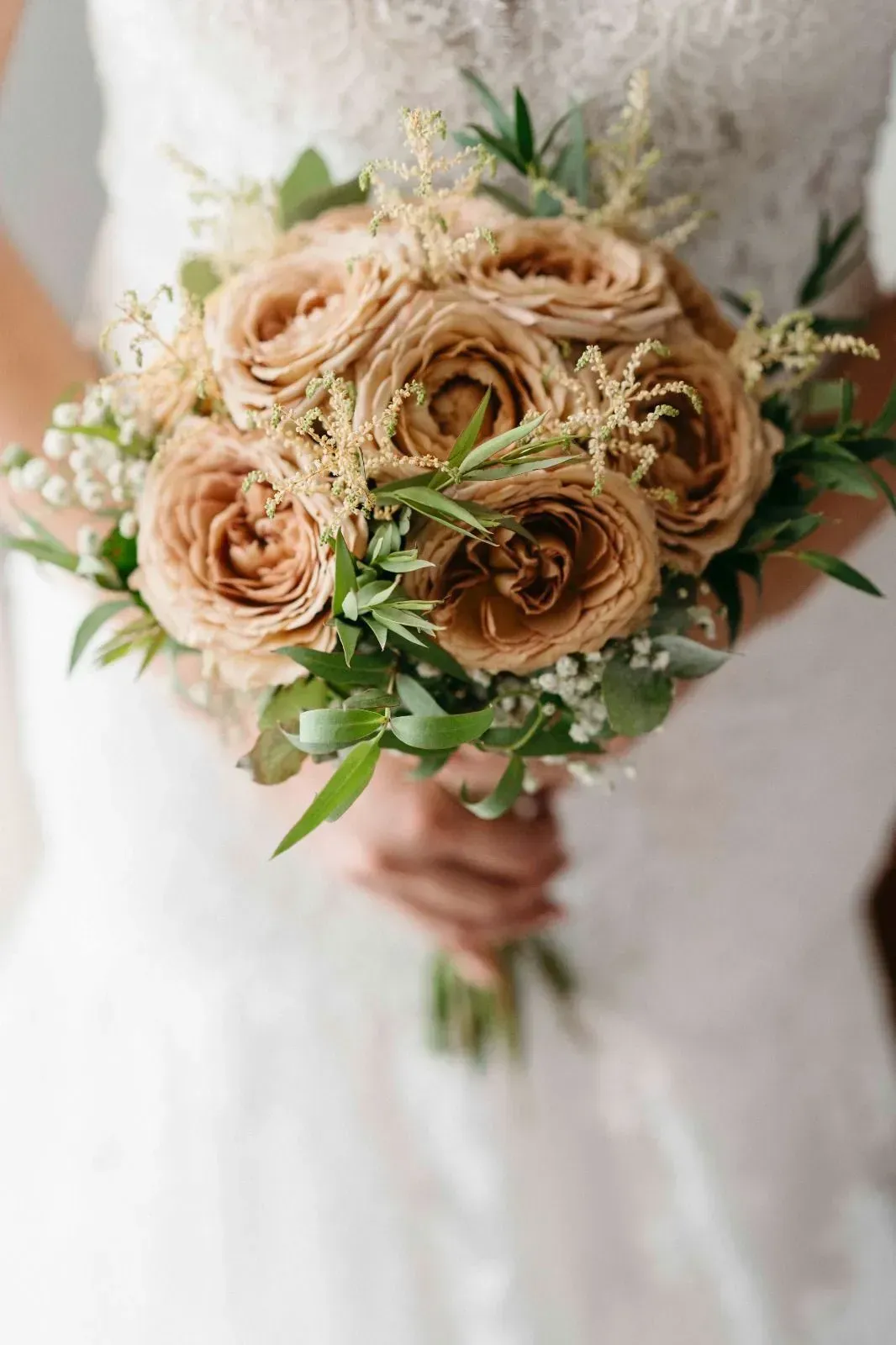Novia sosteniendo un ramo de rosas beige y follaje con un vestido de novia blanco de encaje