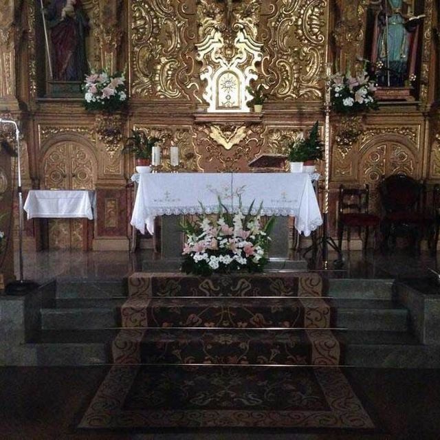 Altar de iglesia con mantel blanco, arreglos florales y un ornamentado fondo dorado sobre una plataforma elevada
