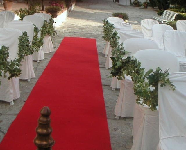 Pasillo de alfombra roja entre filas de sillas blancas decoradas con follaje, preparado para un evento al aire libre.