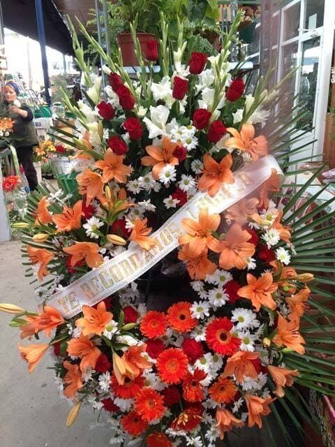 Corona fúnebre colorida de flores rojas, naranjas y blancas, con una cinta blanca sobre un soporte al aire libre