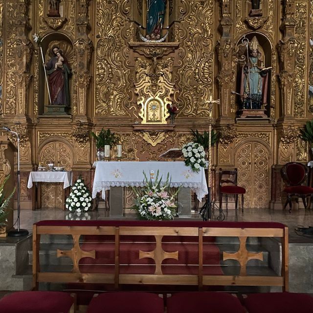 Altar de iglesia ornamentado con manteles blancos, flores, estatuas y un retablo de madera tallada y dorada