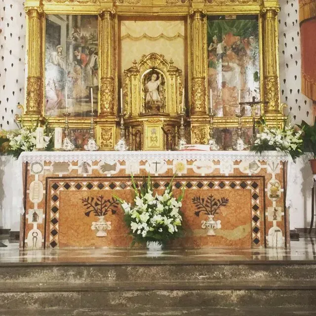 Altar de iglesia dorado y ornamentado, con arreglos florales y paneles decorativos delante de unos escalones