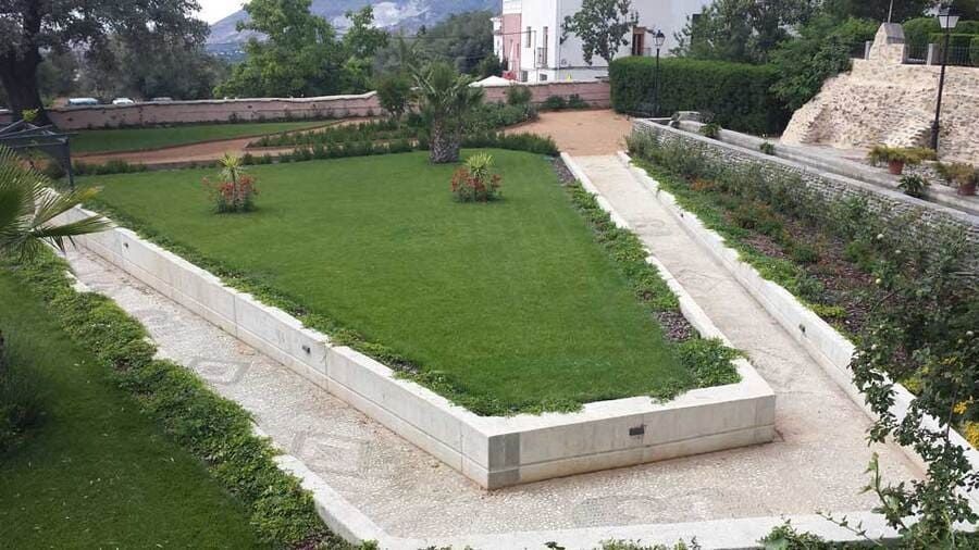 Césped rectangular rodeado de senderos de piedra blanca en una zona ajardinada