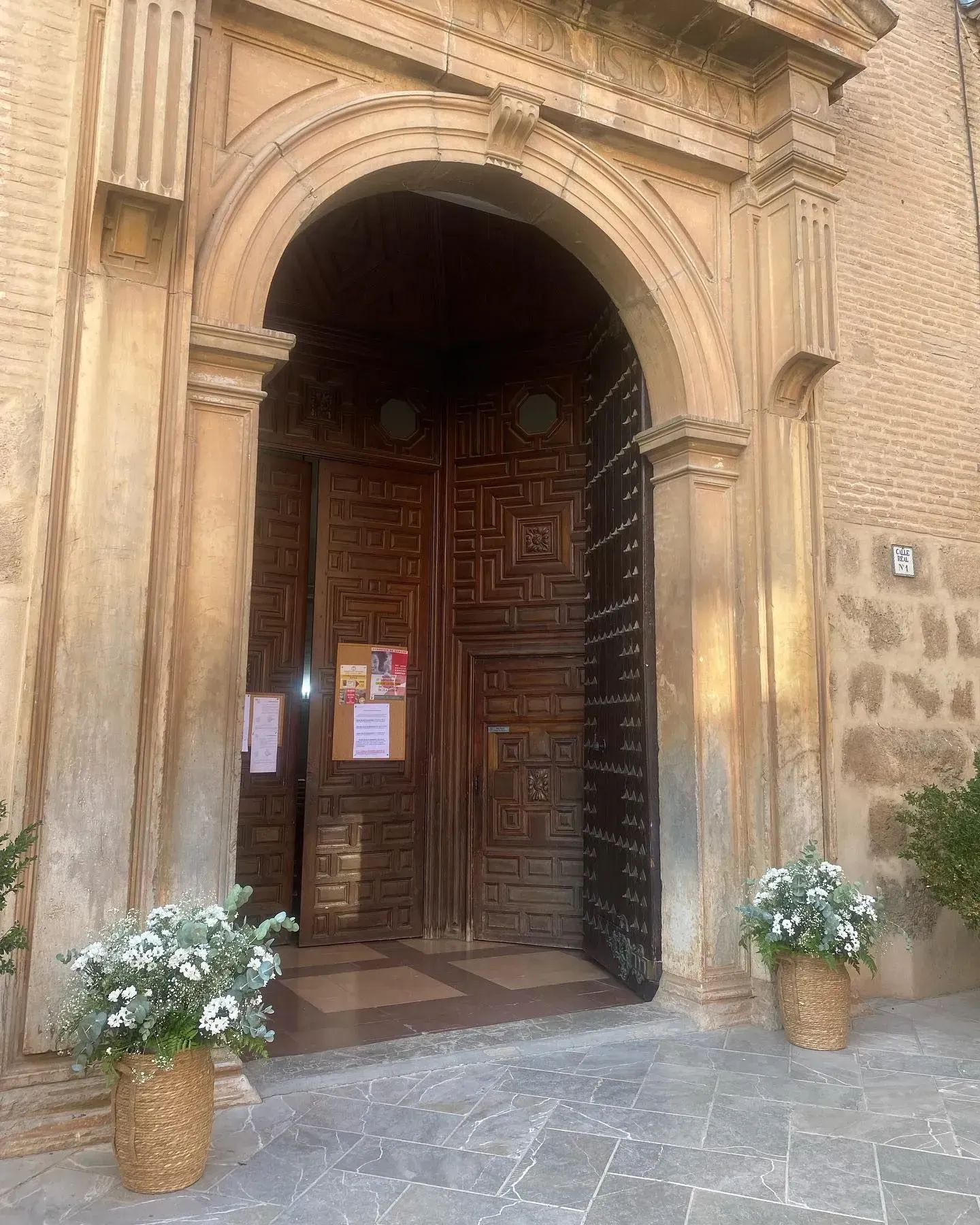 Entrada arqueada ornamentada con puertas de madera tallada y flores blancas en macetas a la entrada