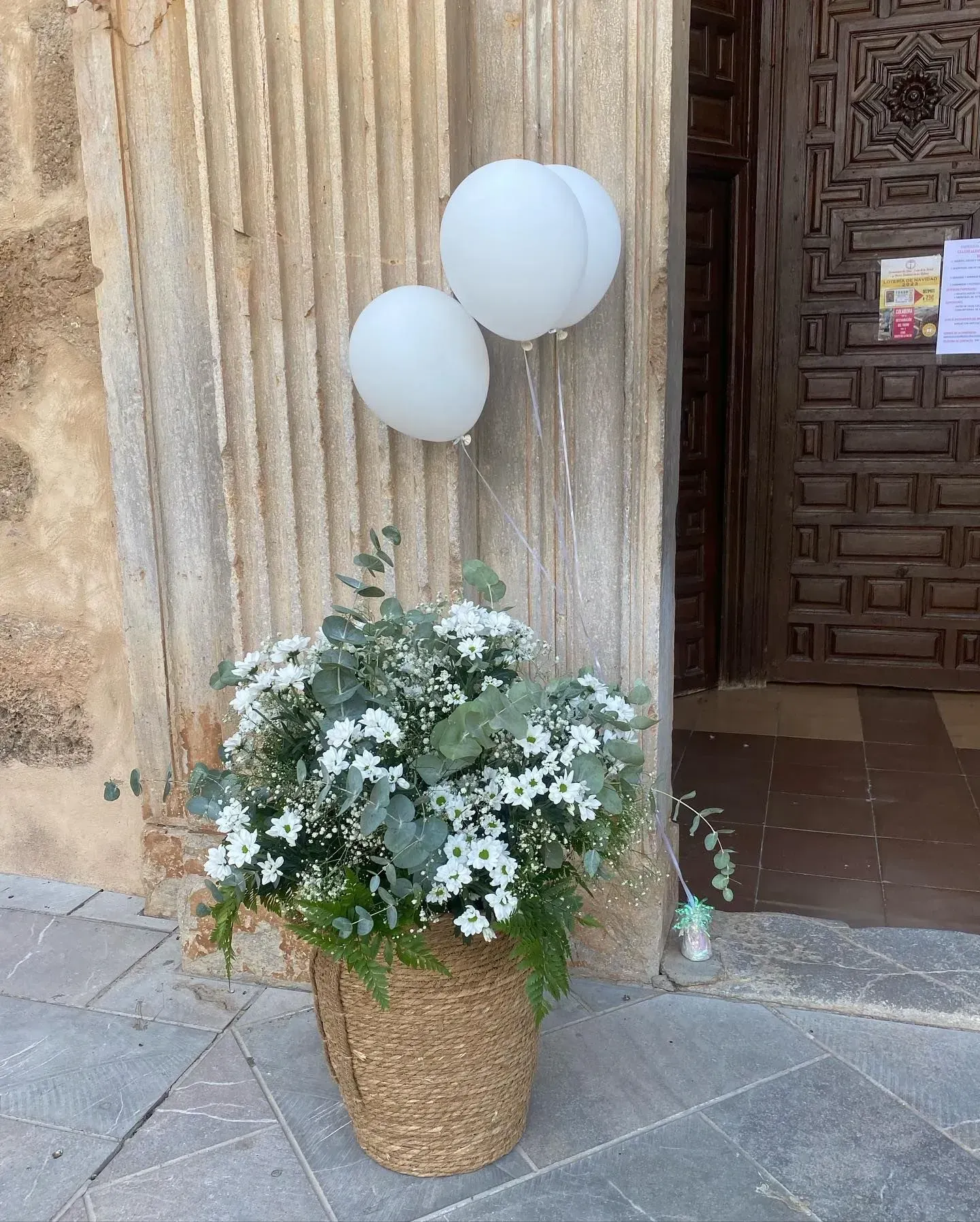 Cesta de mimbre con flores blancas y dos globos blancos junto a una puerta, en un patio de piedra