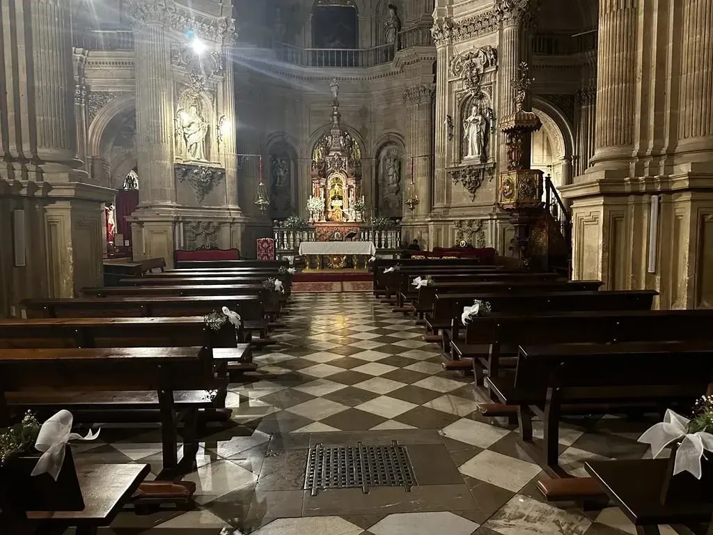 Interior de una gran catedral, con bancos, suelo de cuadros y un altar iluminado al fondo